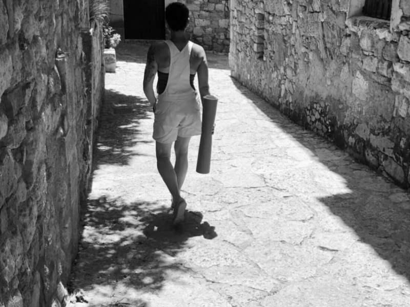 Yoga Frau Müller mit Yogamatte geht barfuß durch schmale Gasse aus Natursteinmauern.