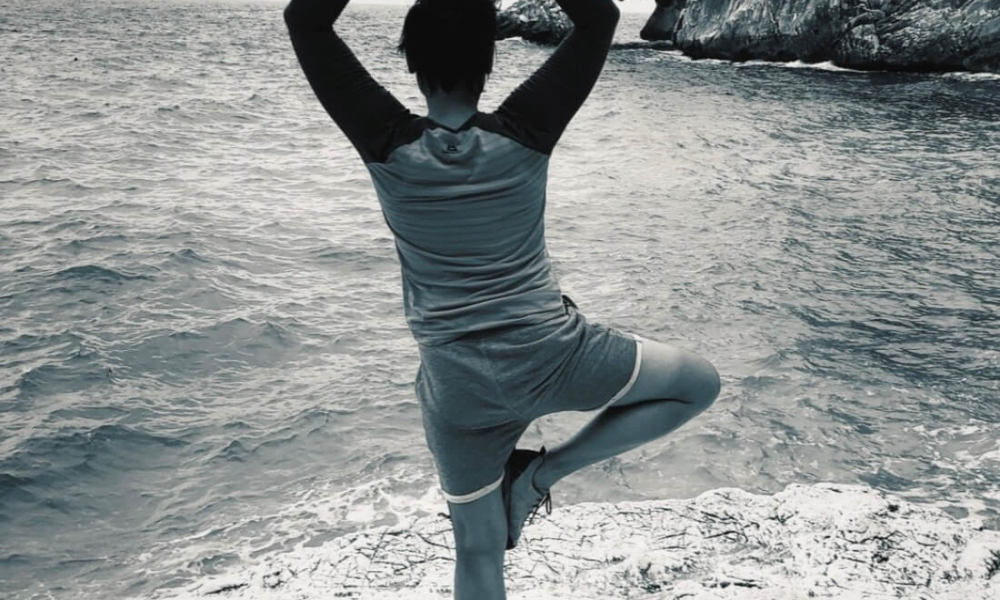 Yoga Frau Müller steht im Baum-Yoga auf Felsen am Meer mit Blick auf Küste und Wasser.