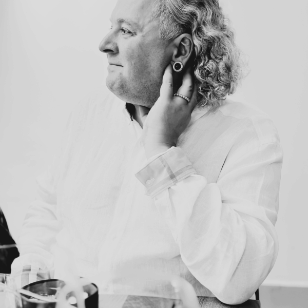 Herr Tribl Herr Tribl mit lockigem Haar und Ohrring in nachdenklicher Pose, sitzt an einem Tisch mit Gläsern, in hellem Hemd.