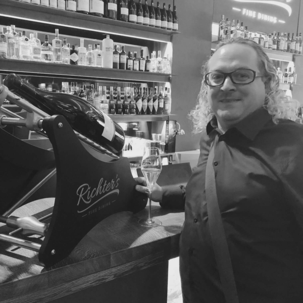 Dekanter Herr Tribl in dunklem Hemd mit Weinglas vor Weinregal bei Richter's Fine Dining, große Weinflasche in Kipphalterung im Vordergrund.