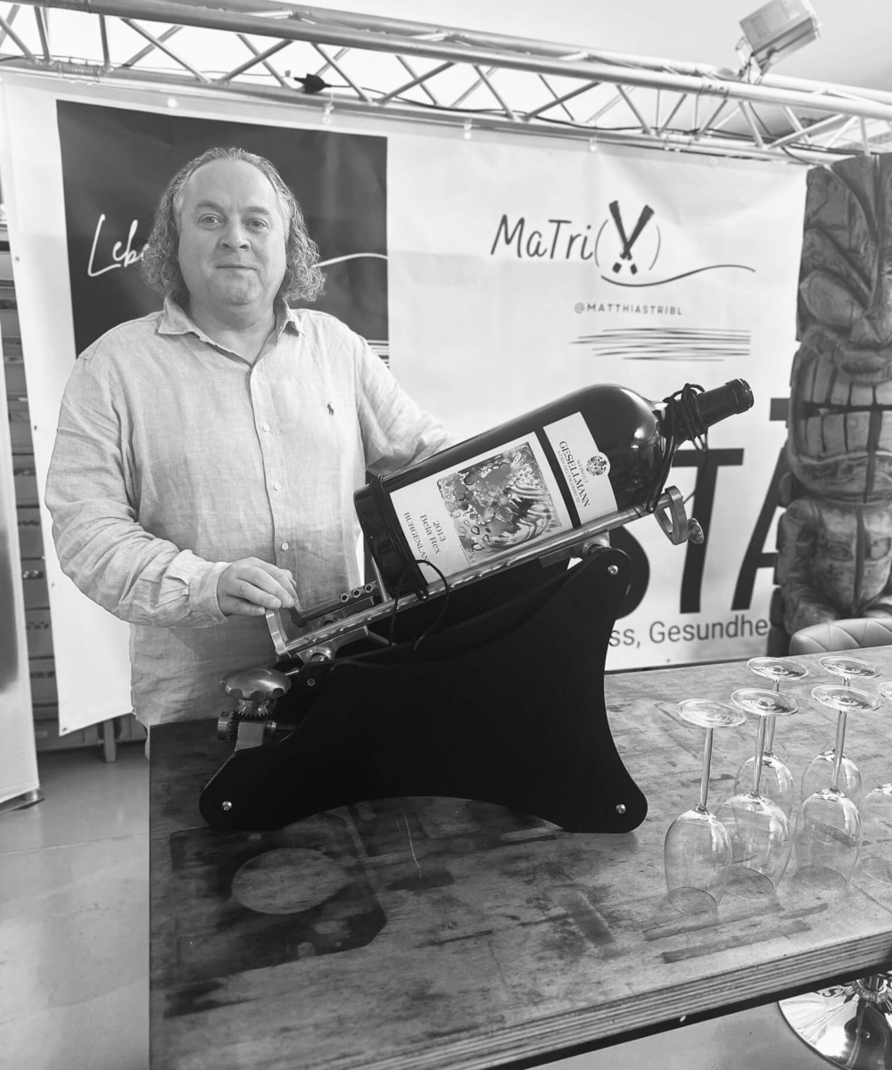 Dekanter Herr Tribl in hellem Hemd präsentiert große Weinflasche in Kipphalterung, umgeben von leeren Weingläsern, vor Werbebannern mit 'MaTriX' und 'Lebensfreude' Schriftzügen.